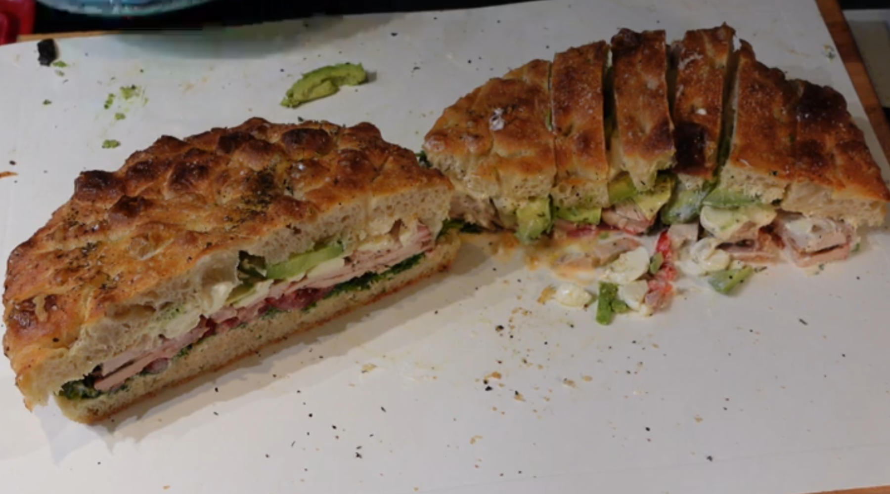 Two halves of a thick focaccia sandwich filled with meats, cheese, avocado, and vegetables on a white cutting board.