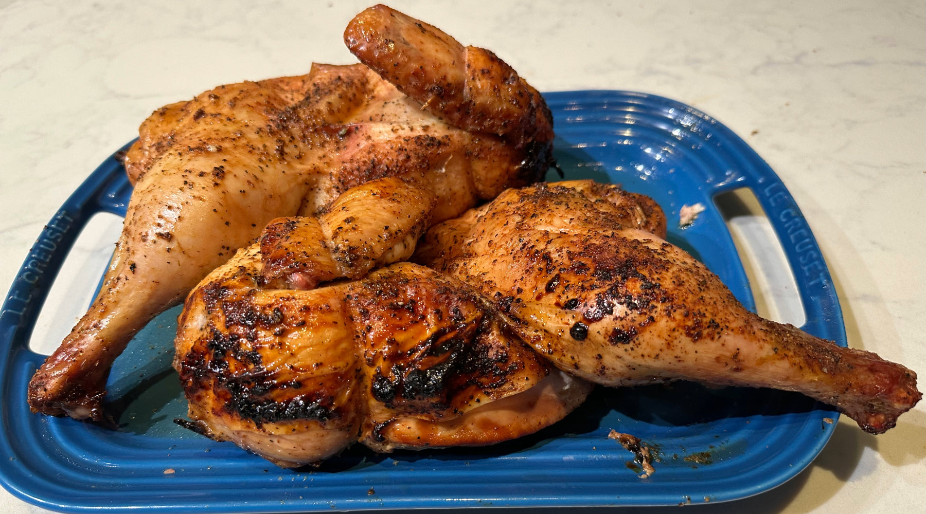 Grilled and seasoned split chicken halves served on a blue Le Creuset tray, ready to be carved and enjoyed.