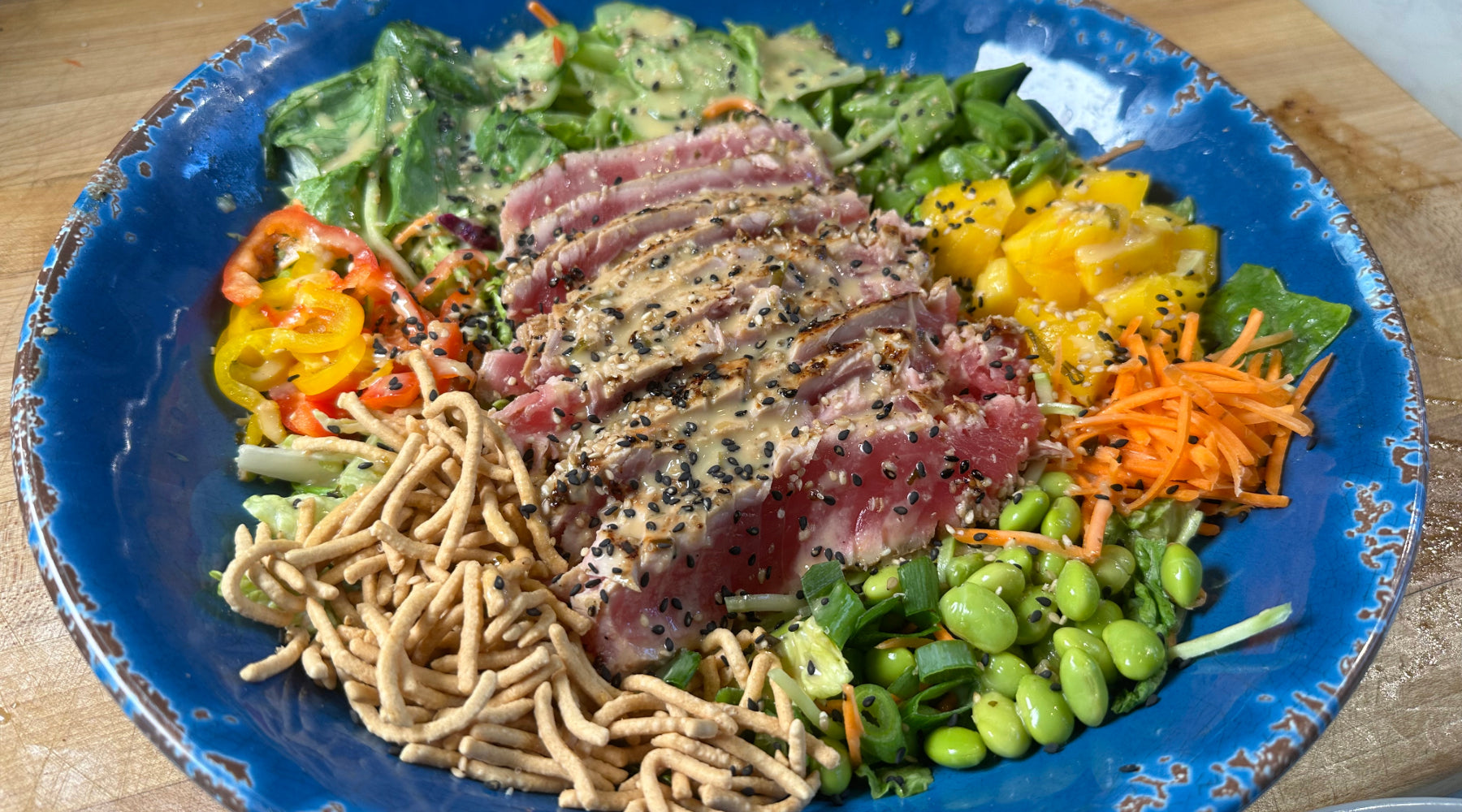 Seared Ahi Tuna Asian Cobb Salad with shredded veggies, mango, edamame, and sesame-ginger dressing in a blue bowl.