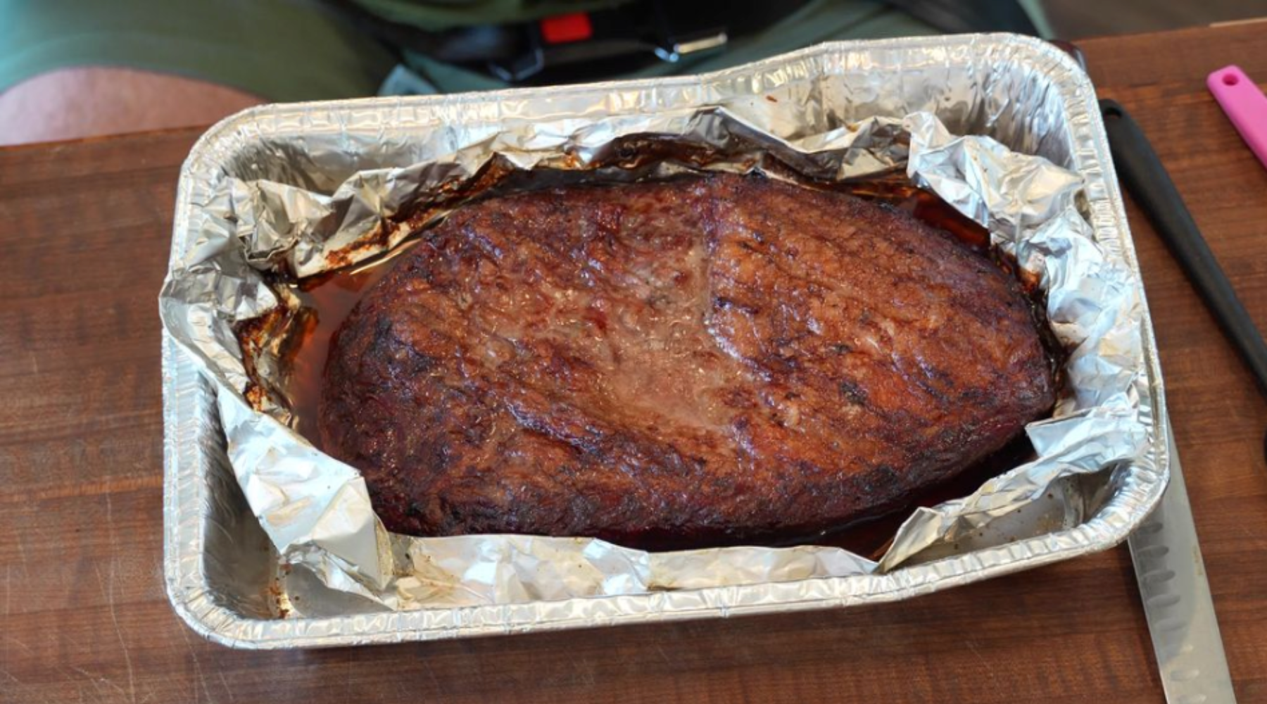 Large smoked brisket resting in a foil-lined aluminum tray, showing a dark, caramelized crust.