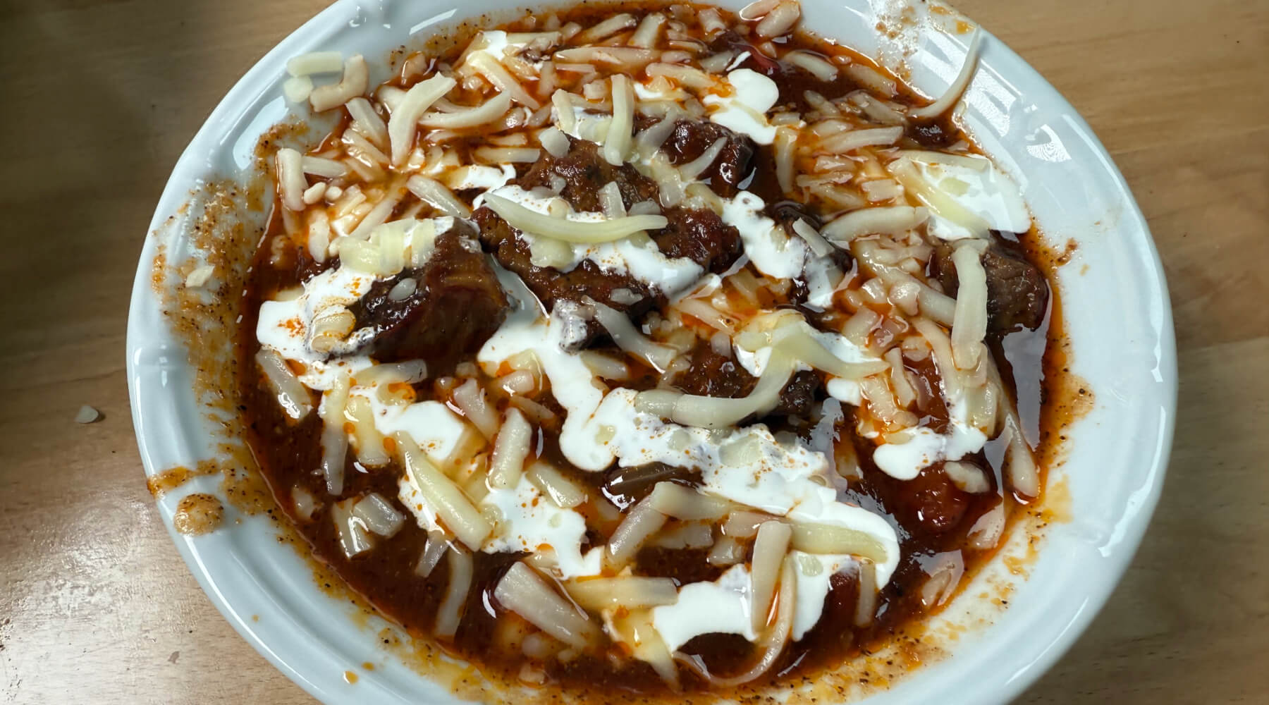 Bowl of chili topped with shredded cheese and sour cream, served in a white dish with some sauce spilled on the rim.