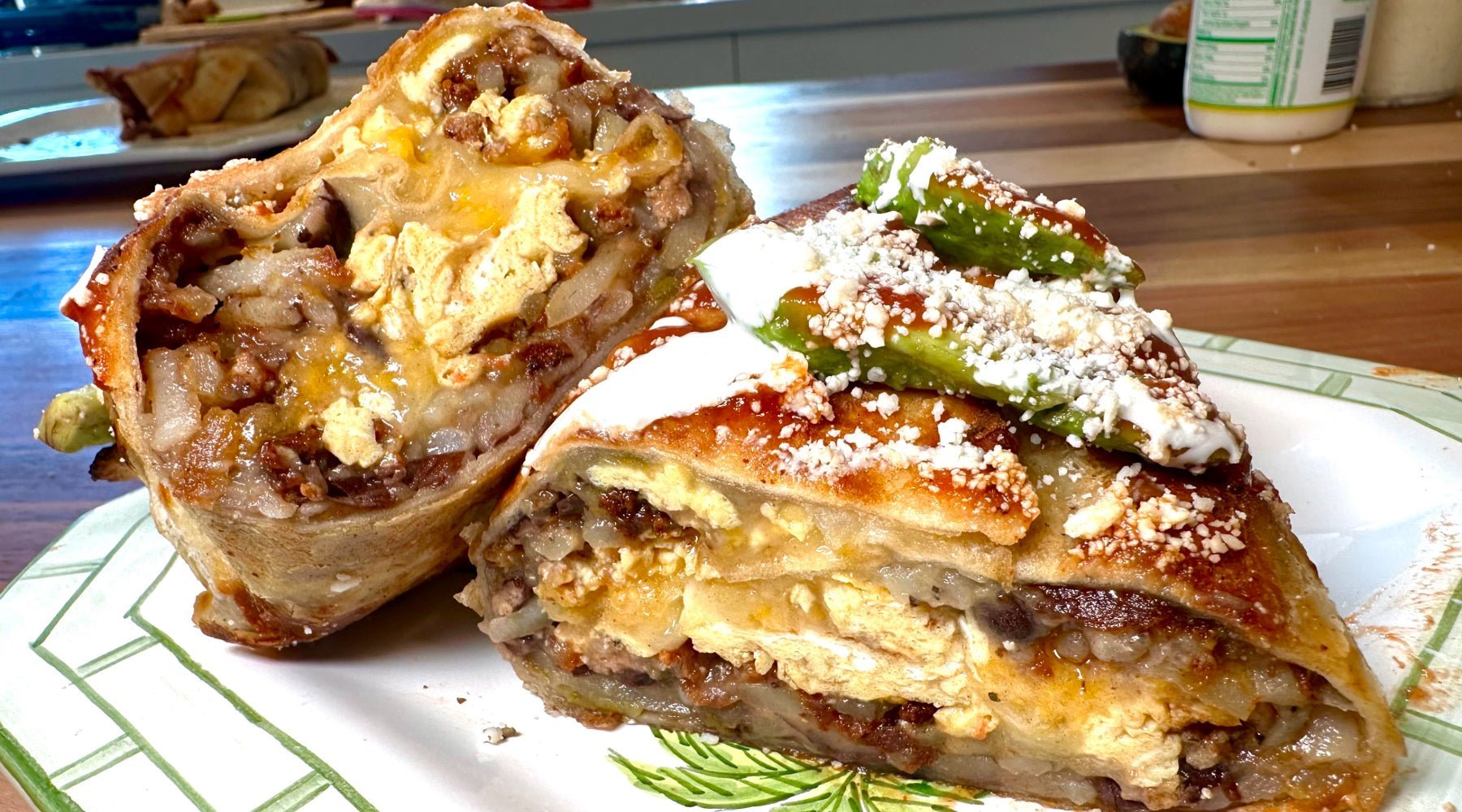 A large burrito cut in half, revealing layers of scrambled eggs, ground meat, melted cheese, potatoes, and beans. The burrito is topped with avocado slices, crumbled cheese, and a drizzle of sauce.