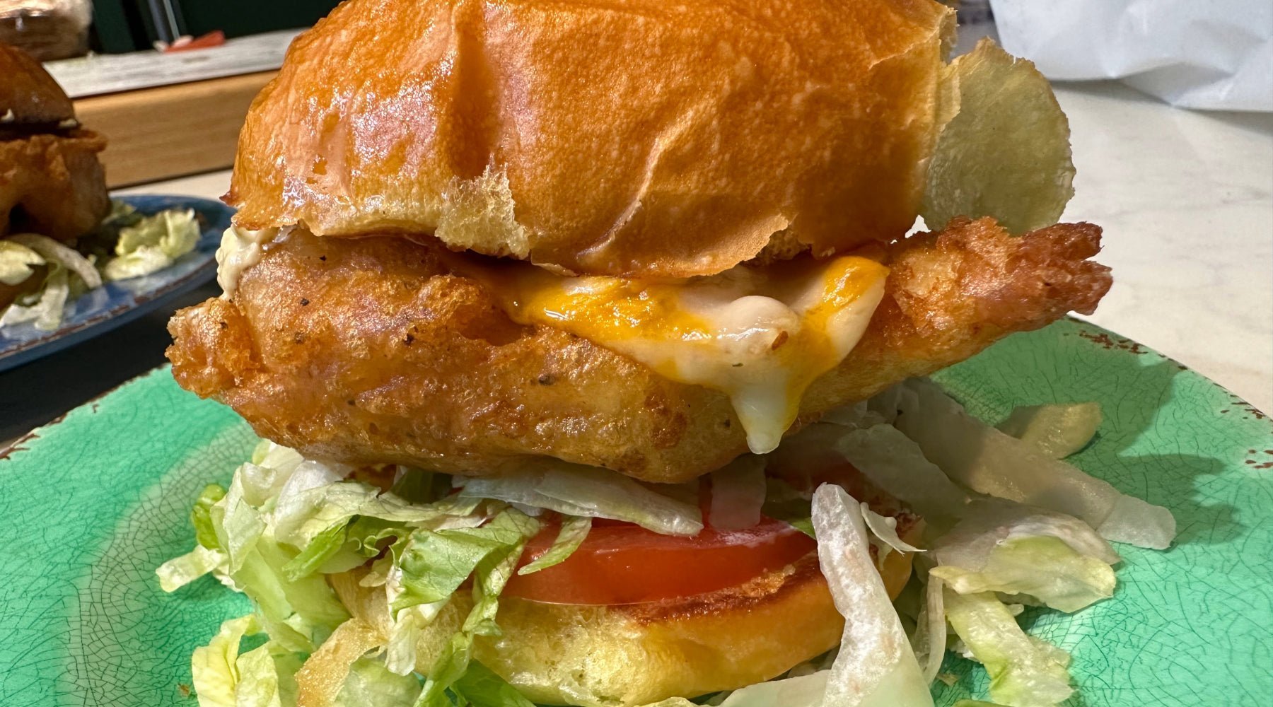 A crispy fried chicken sandwich topped with melted cheese, lettuce, and a slice of tomato, served on a golden, toasted bun.