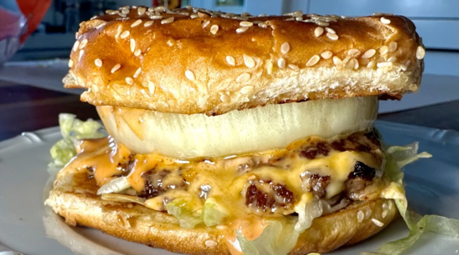 A close-up of a juicy cheeseburger on a white plate. The burger has a sesame seed bun, a thick slice of onion, melted cheese, shredded lettuce, and a beef patty with sauce dripping down the sides.