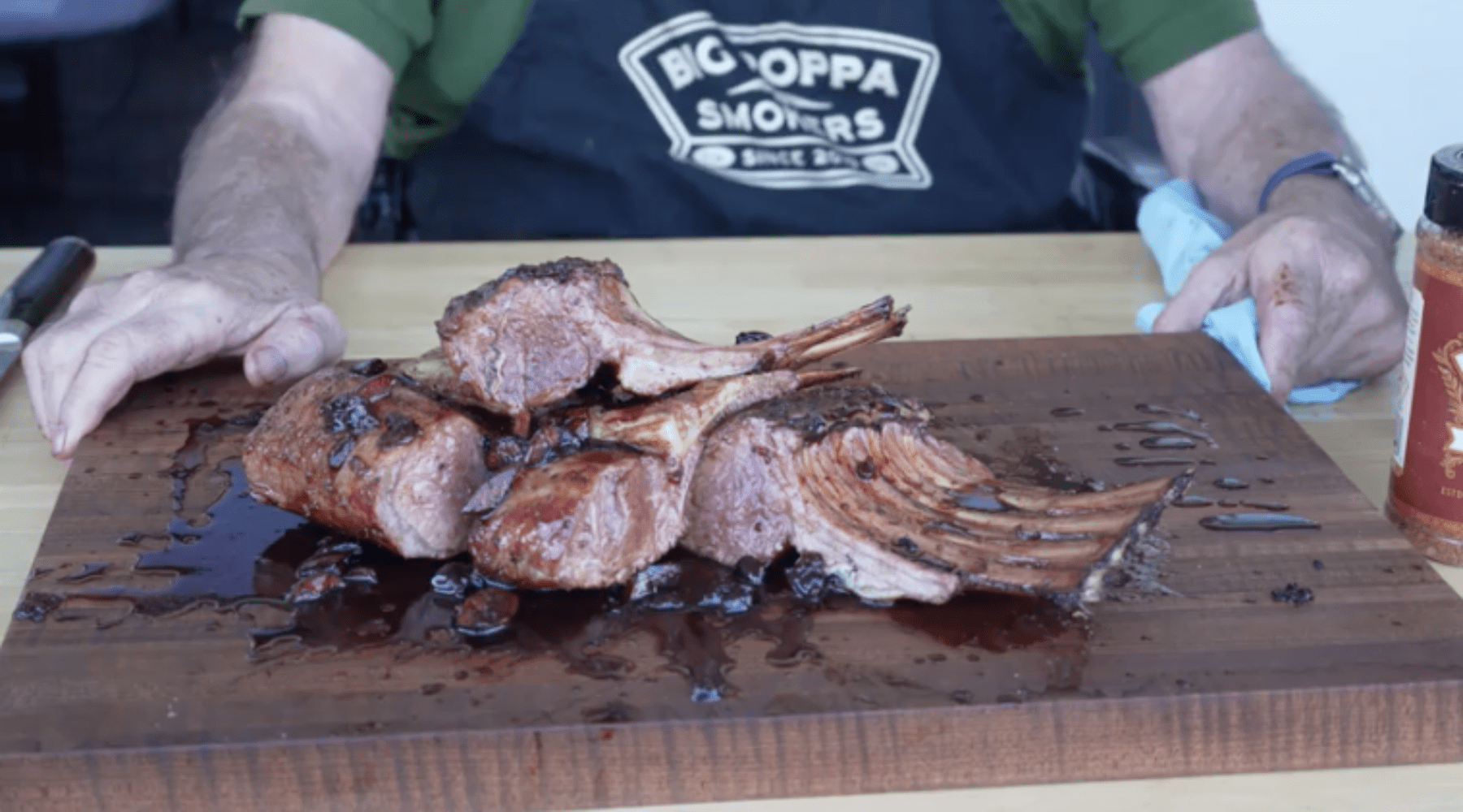 Grilled Cheraki lamb chops resting on a wooden cutting board, glistening with juices beside a person in a black apron.