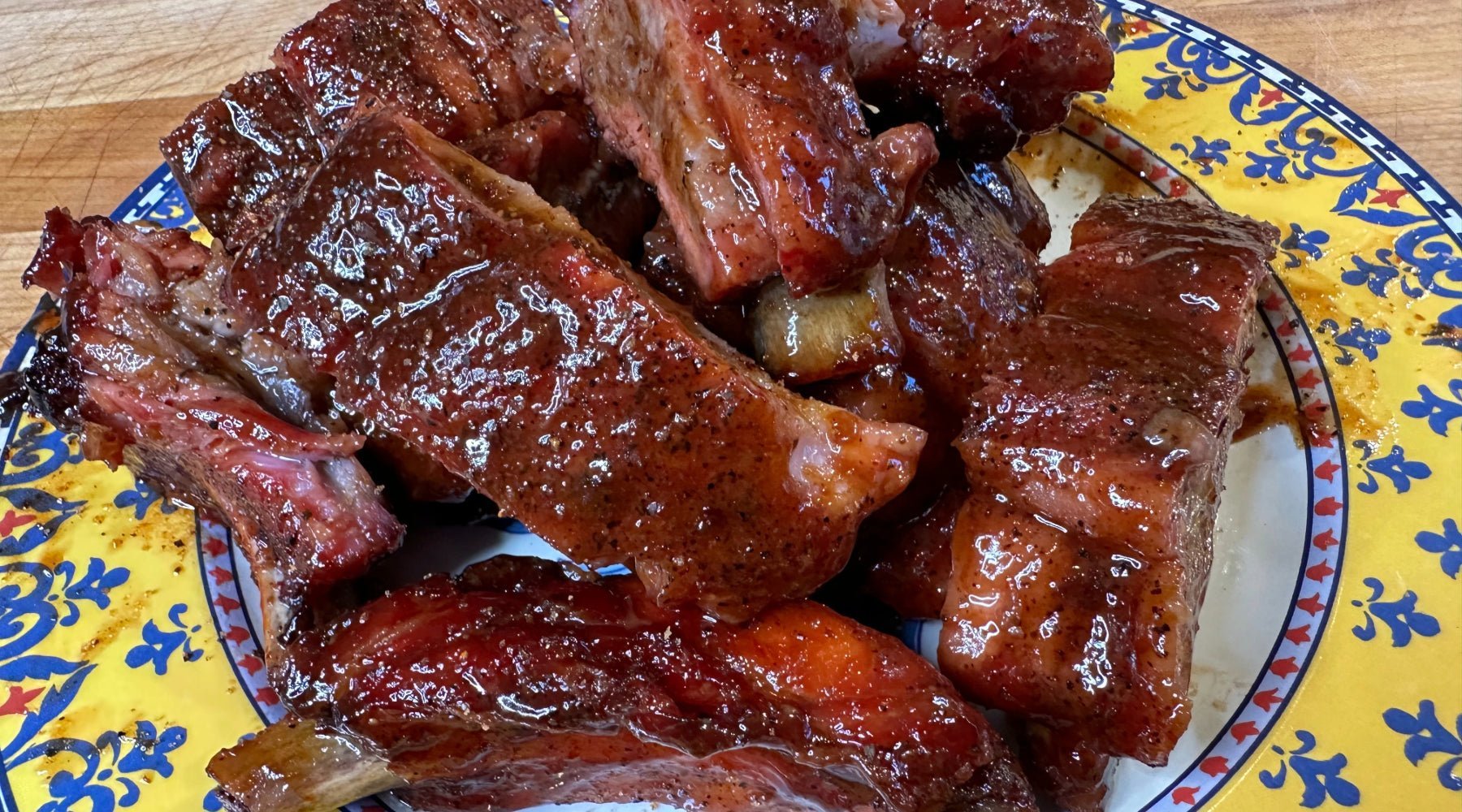 A plate of glazed, barbecued ribs piled high on a colorful, patterned plate. The ribs are coated in a shiny, rich sauce, highlighting the charred and juicy meat underneath.