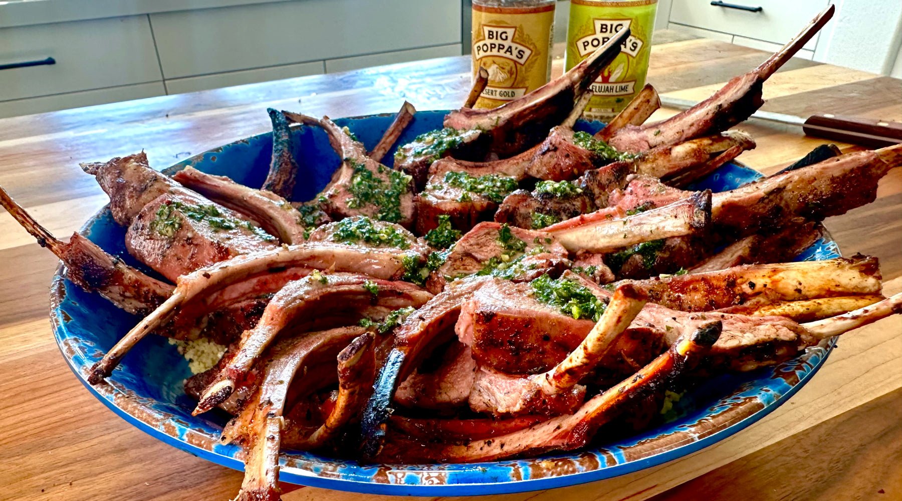 A large blue platter filled with grilled lamb chops topped with green herb sauce, with Big Poppa’s seasoning bottles in the background.