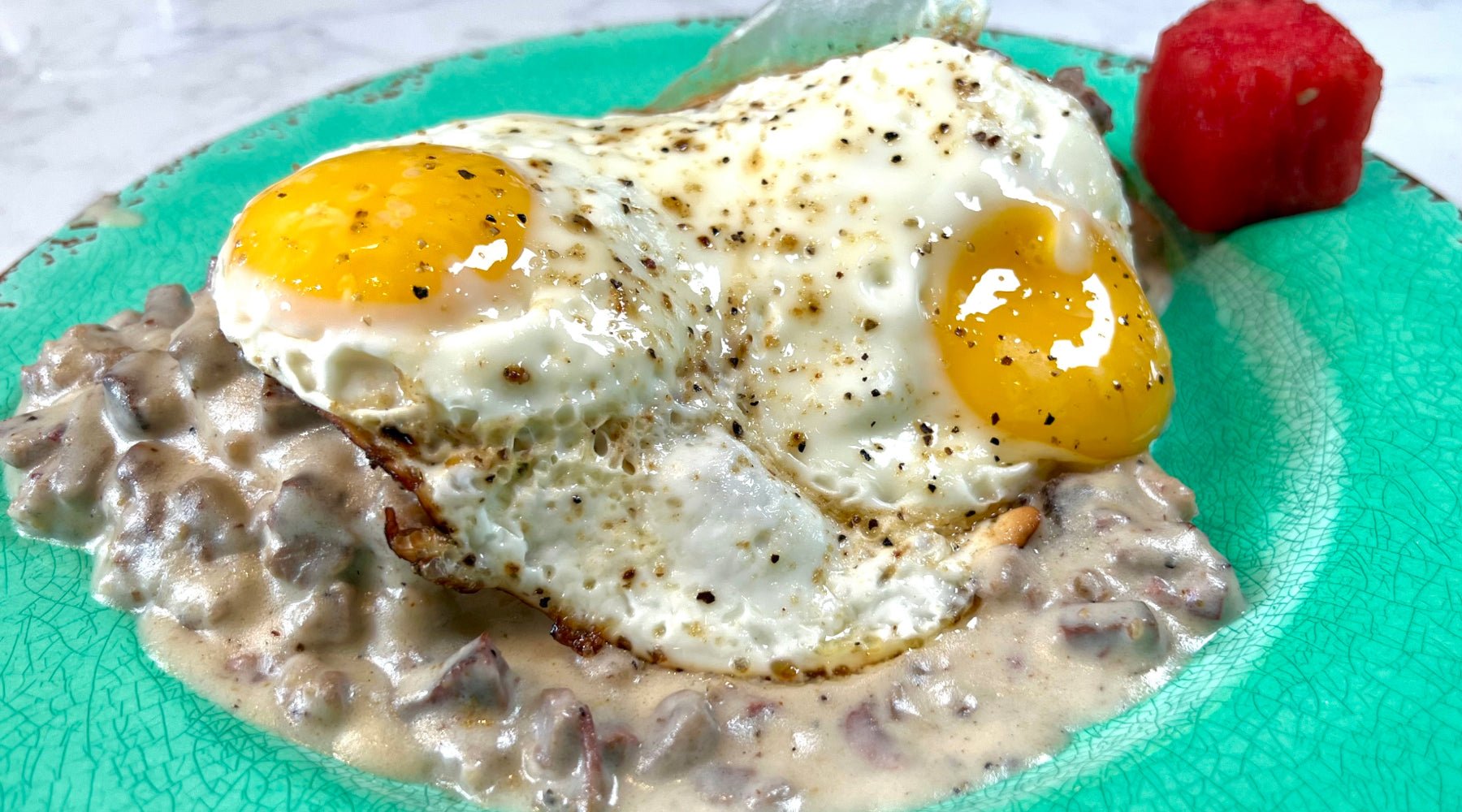 Two sunny-side-up eggs over sausage gravy on a biscuit, served on a turquoise plate with a small piece of watermelon.