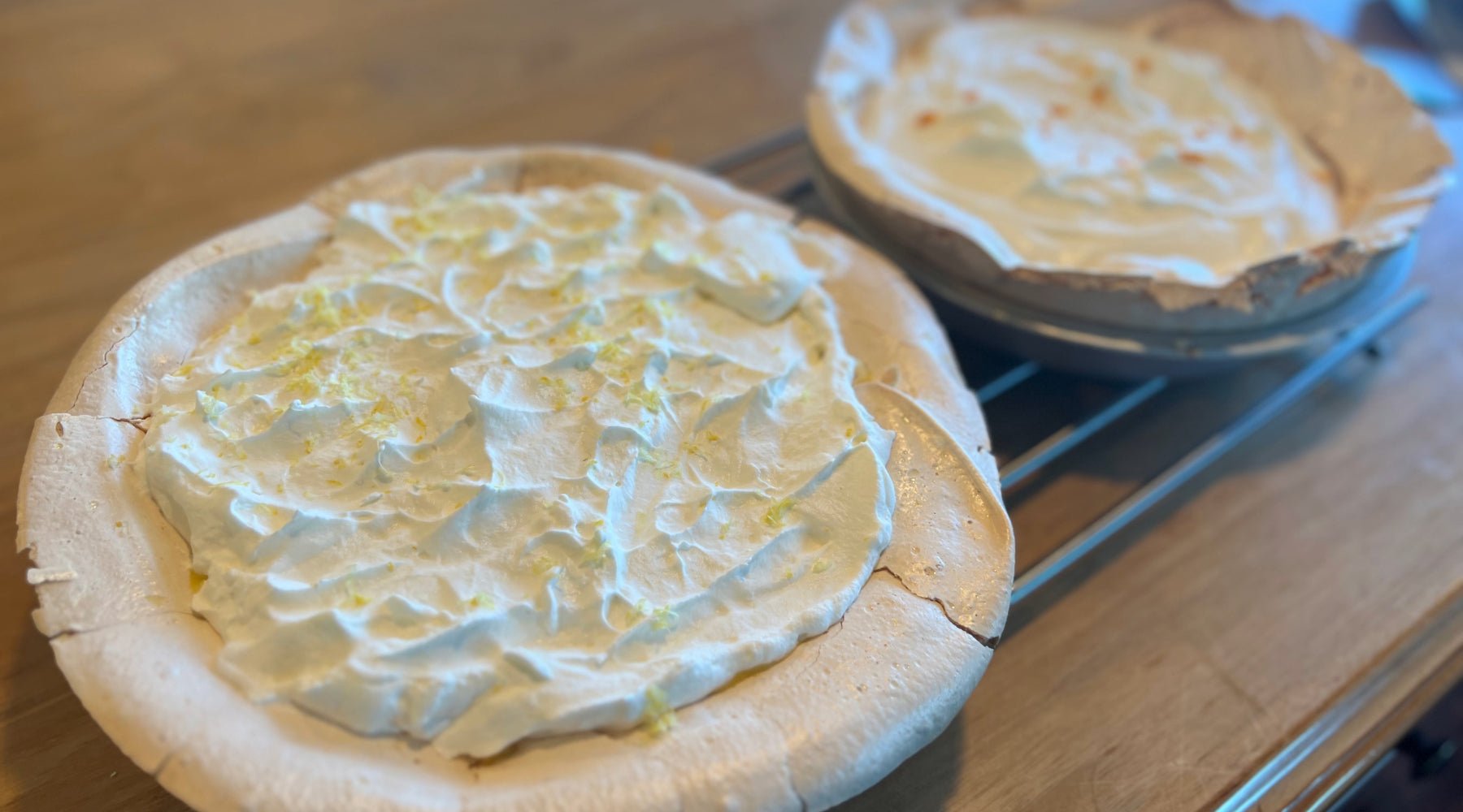 Two lemon meringue pies with fluffy, golden-tipped meringue tops cooling on a wire rack.