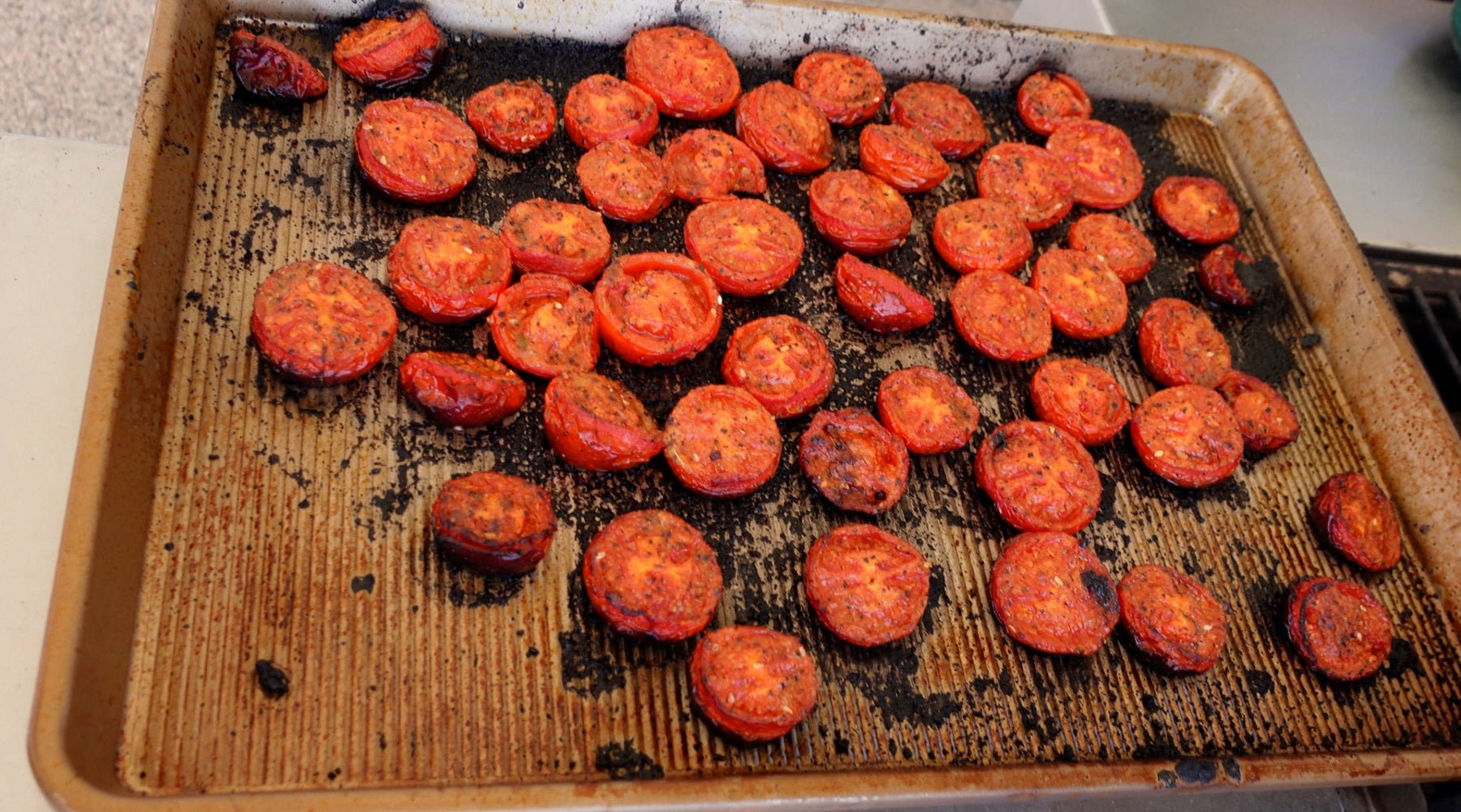 Roasted tomato halves on a well-used baking sheet, some slightly charred, arranged in a loose, scattered pattern.