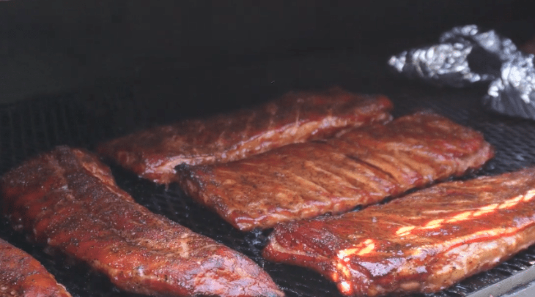 Glazed pork ribs smoking on a grill, nearly done cooking, with foil-wrapped items in the background.