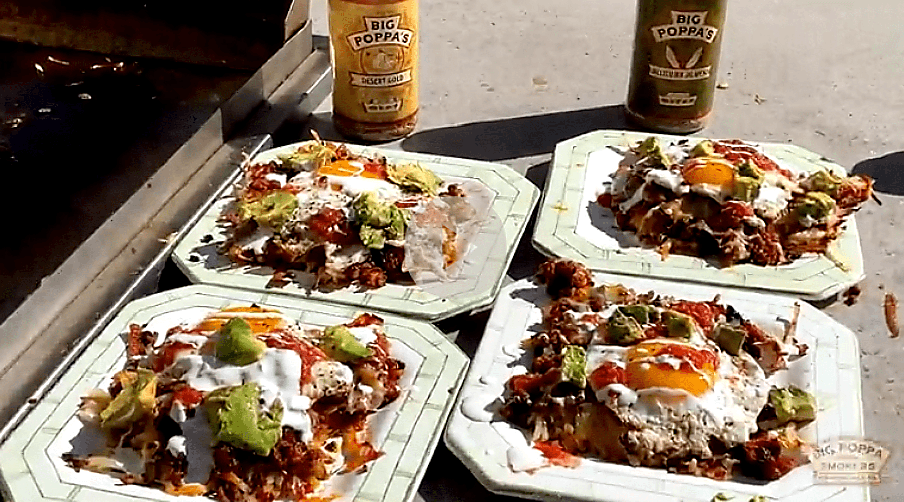 Four plates of loaded hash topped with eggs, guacamole, sour cream, and salsa beside Big Poppa’s seasoning bottles on an outdoor grill surface.