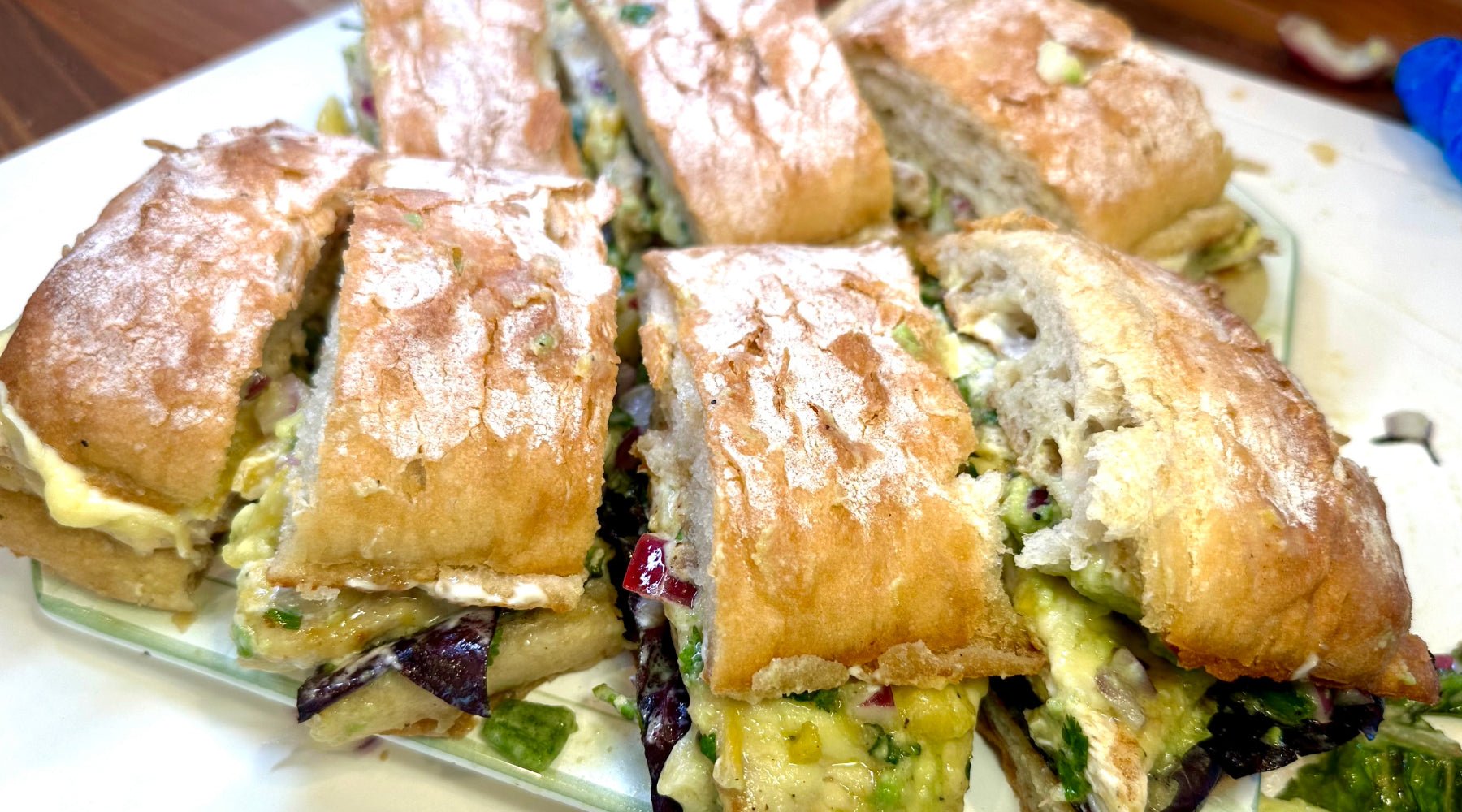 Sliced sandwiches on crusty bread, filled with a creamy mixture of vegetables, cheese, and greens, served on a glass plate.