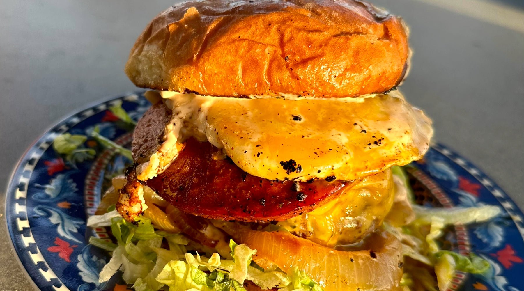 A close-up of a burger with melted cheese, a fried egg, crispy bologna, grilled onions, and lettuce on a toasted bun, served on a plate.