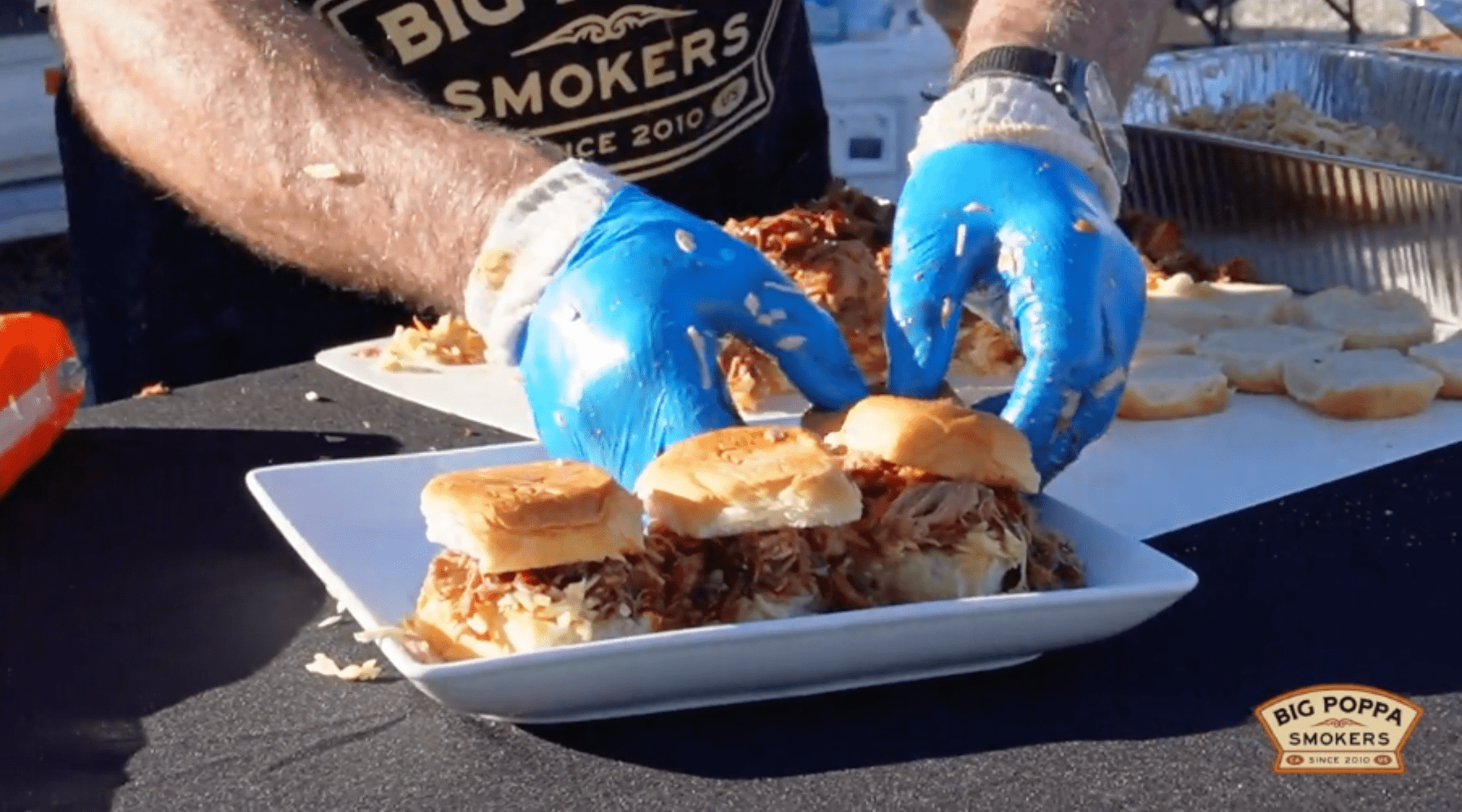 A person wearing blue gloves assembles pulled pork sliders on a white plate at an outdoor event.