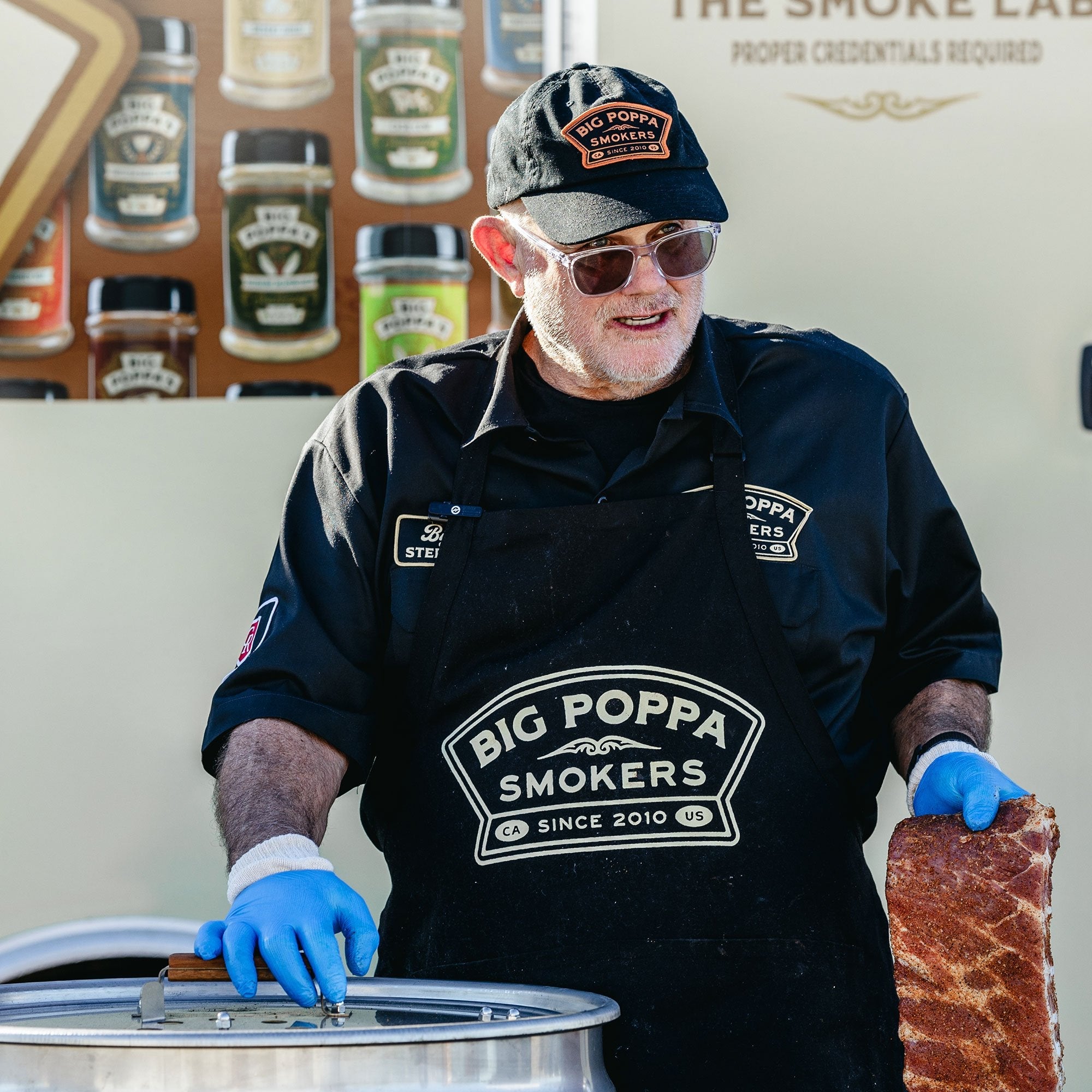 Big Poppa showing off his BBQ merch, wearing a branded apron, shirt, and hat.