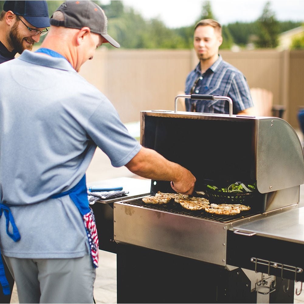 Man in blue shirt grills meat & veggies on MAK Grill while two others watch, all outdoors on a patio in a casual BBQ setting