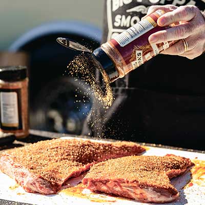 Big Poppa sprinkling Double Secret Steak Seasoning on two tri-tips pieces of meat that are generously coated.