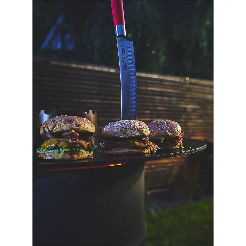 Red-handled slicing knife stuck upright in a griddle beside three grilled burgers outdoors at night.