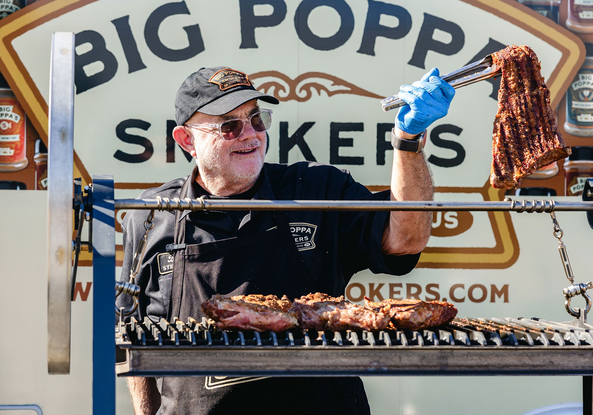 Sterling "Big Poppa" Ball holding up a tri-tip with tongs, as he is getting ready to flip on the Santa Maria Smoker.