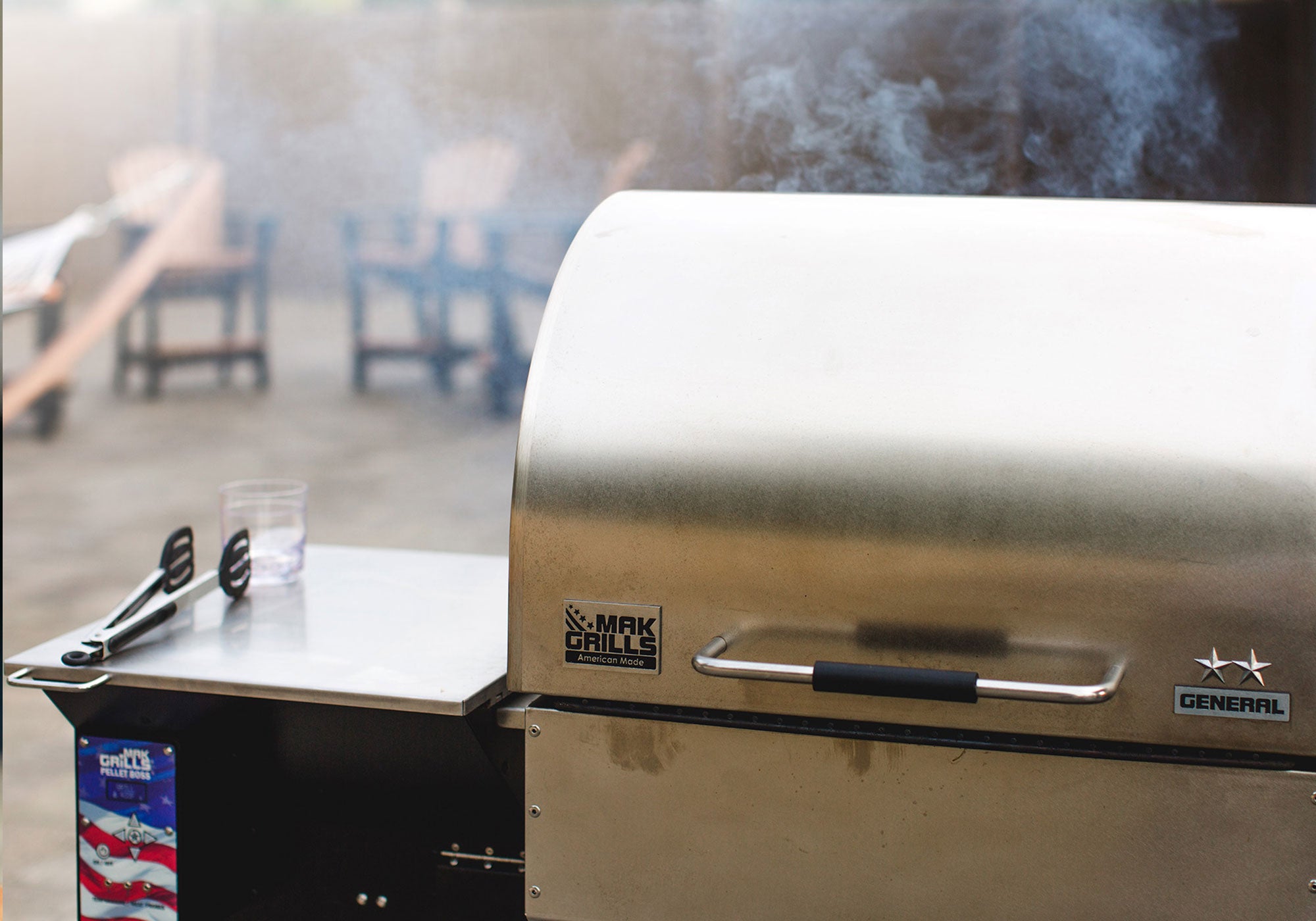 Image of a MAK 2 Star Grill that has meat on it cooking and smoke coming out. In the distance you can see chairs.