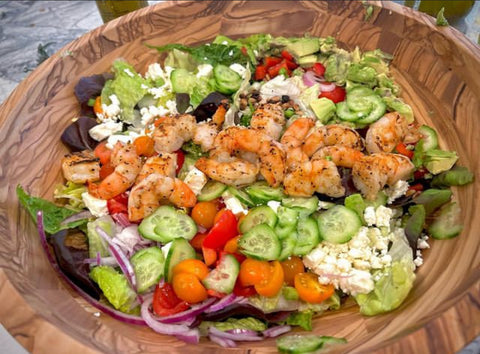 A colorful salad featuring grilled shrimp on top, with mixed greens, cherry tomatoes, cucumbers, red onions, and crumbled cheese in a large wooden bowl.