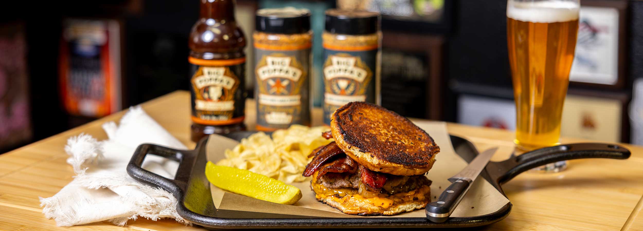 A grilled sandwich with bacon and melted cheese, served with chips and pickle, displayed in front of BBQ sauces and seasonings.