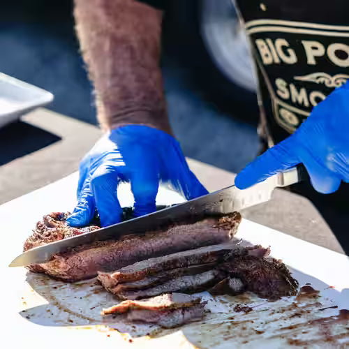 Person cutting meat with a knife and blue gloves on a white surface.