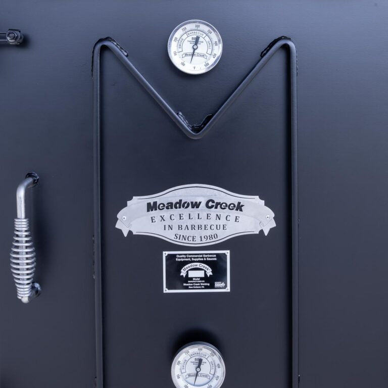 Close-up of the thermometer and logo on the upper door of the Meadow Creek BX50 smoker, displaying the text 'Meadow Creek Excellence in Barbecue Since 1980' above the temperature gauge.