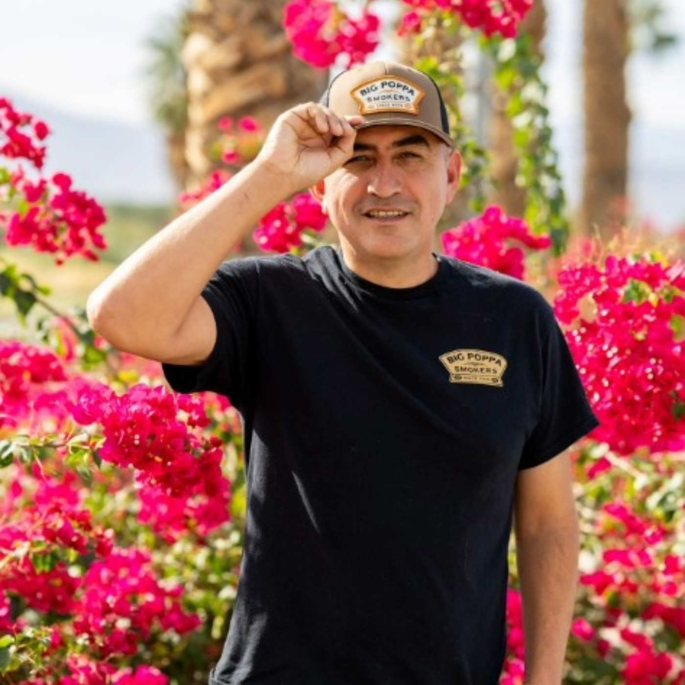 Man wearing a black t-shirt with a logo and a cap, standing in front of pink flowers.