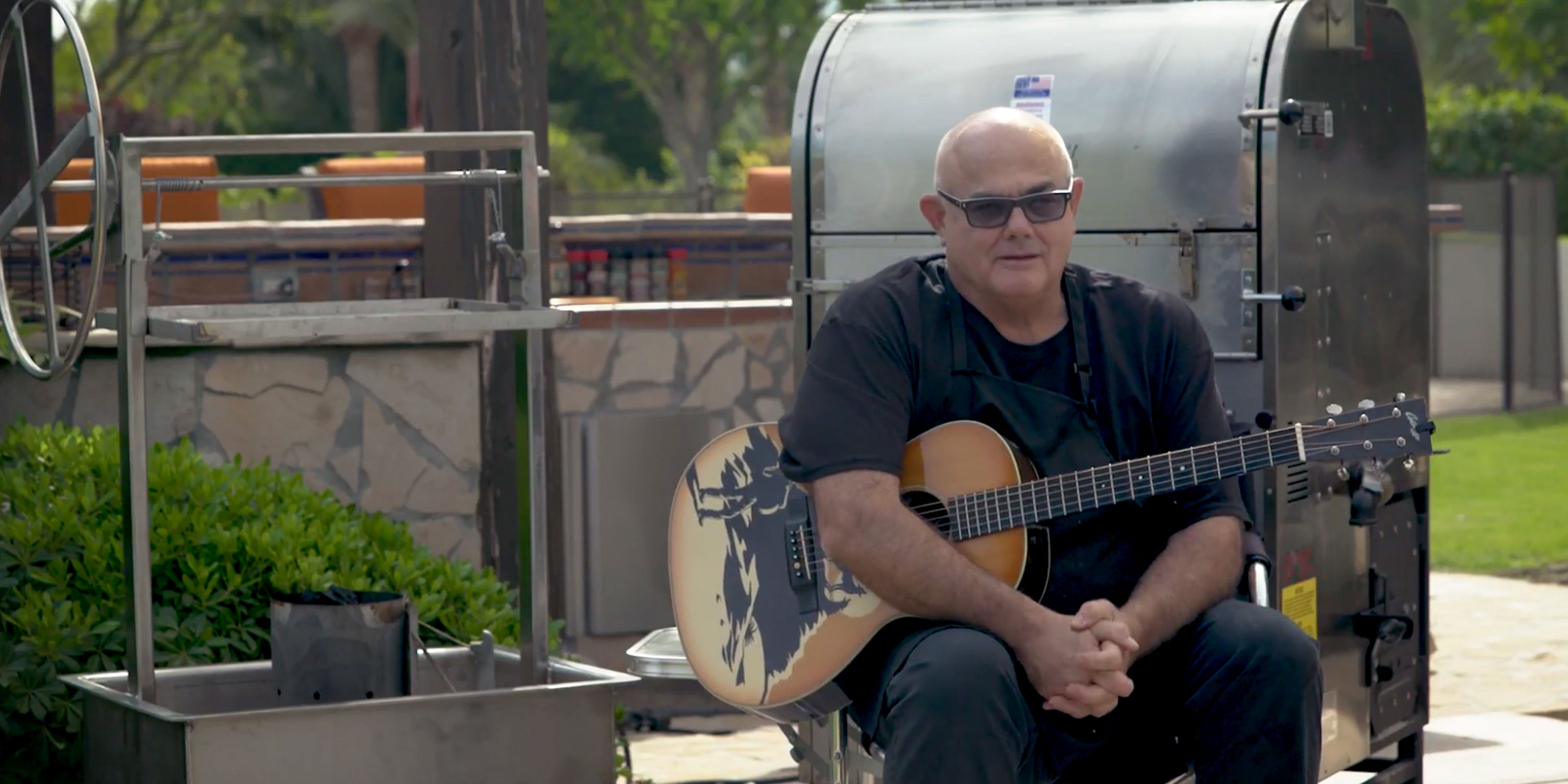 Sterling "Big Poppa" Ball sitting outside in front of his smokers with his guitar.