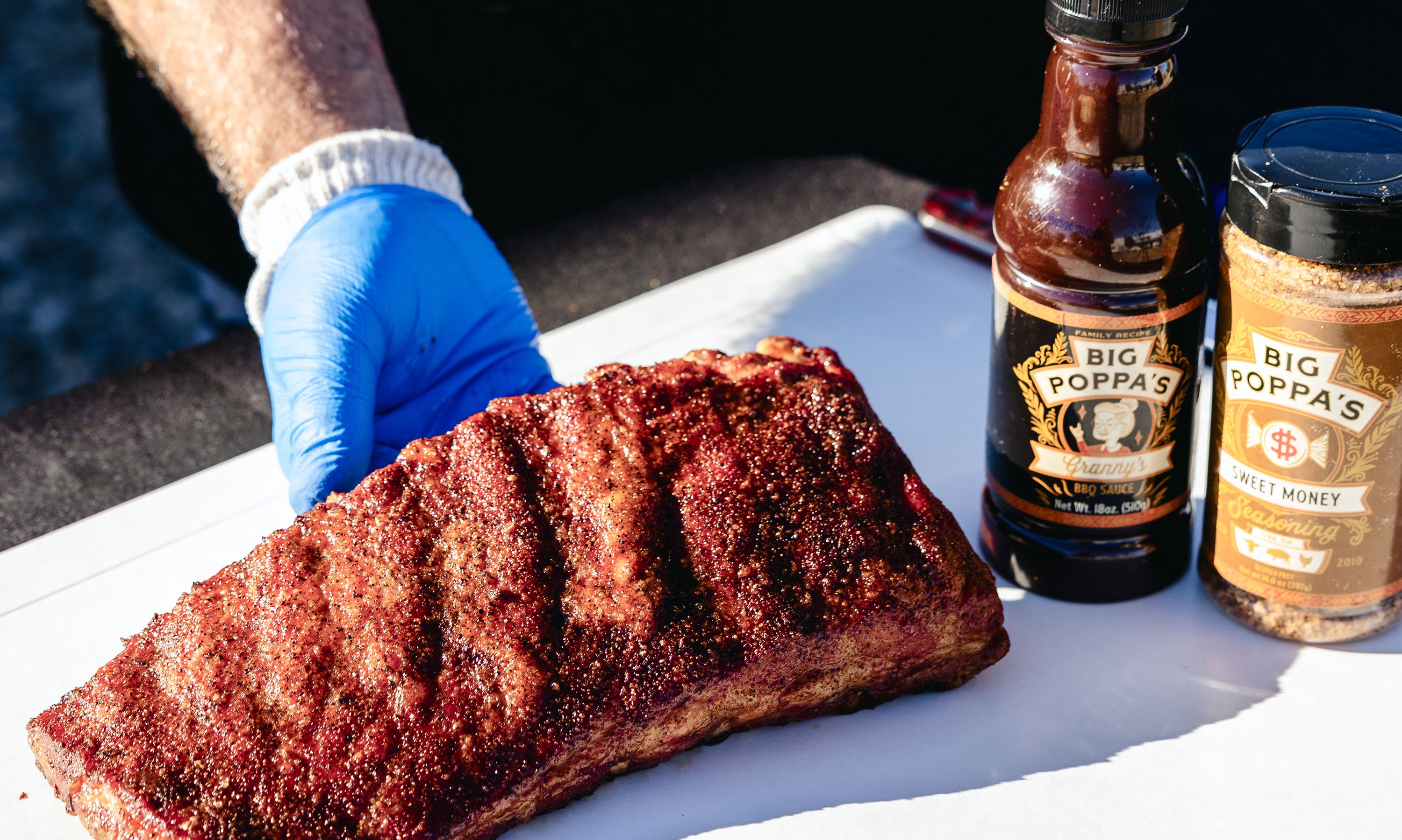 A succulent slab of ribs that is getting ready for a generously coated with Big Poppa's Granny's BBQ Sauce and sprinkled with Sweet Money BBQ Rub, creating a glossy and richly seasoned surface. The ribs are perfectly cooked, showcasing a deep, caramelized crust that highlights the savory and slightly sweet flavors of the seasoning.