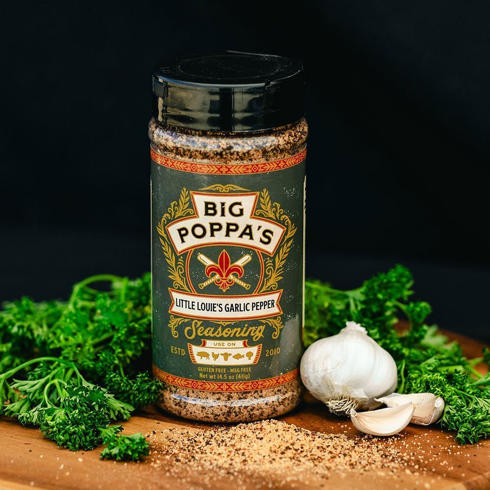 A jar of Big Poppa's Little Louie's Garlic Pepper Seasoning placed on a wooden board surrounded by fresh parsley and whole garlic bulbs. The seasoning jar has a black lid and a decorative label, and is displayed against a dark background.