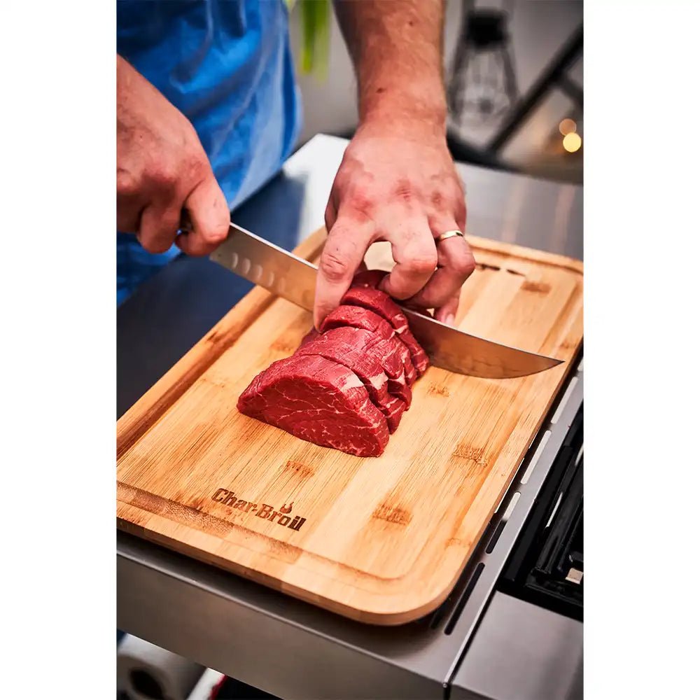 Person slicing raw beef tenderloin on a wooden Char-Broil cutting board with a chef’s knife.