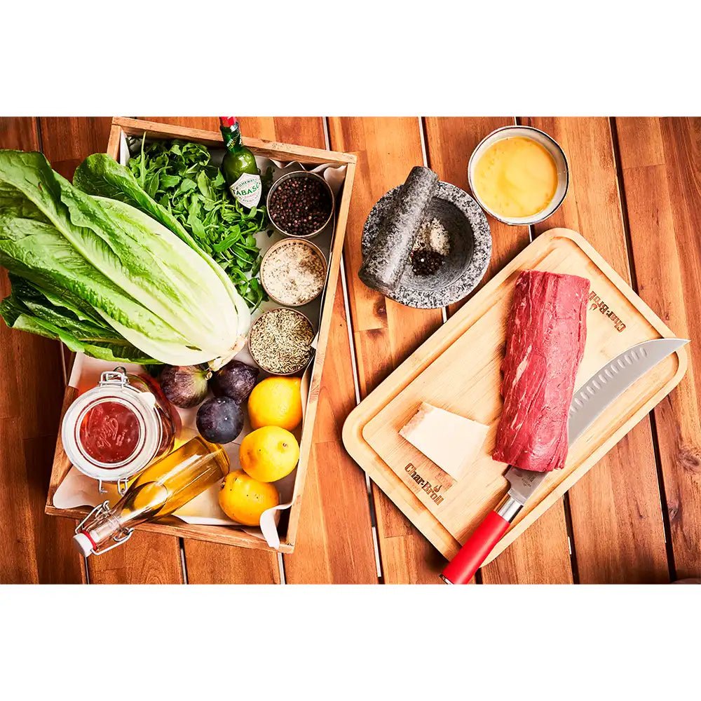 Top view of cutting board with raw beef, cheese wedge, red-handled knife, and fresh produce in a box.