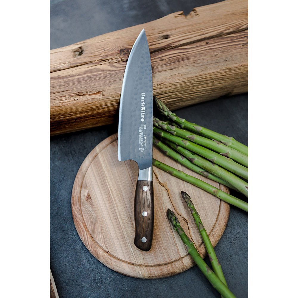 DarkNitro chef's knife with hammered blade on round wood board beside asparagus, set against a rustic wooden log background.
