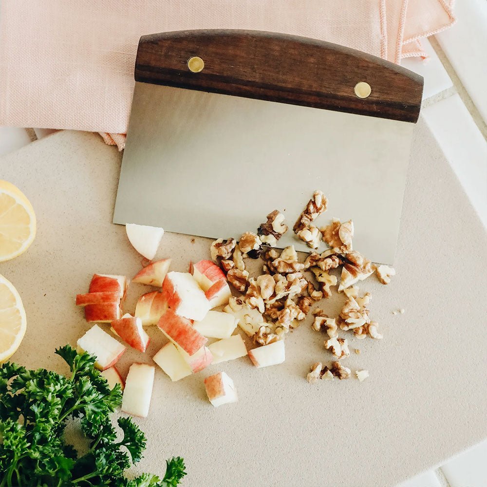 Dough scraper on cutting board with chopped apples, walnuts, parsley, and lemon.