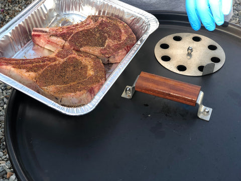 Raw thick-cut steaks in a disposable aluminum tray, seasoned with a dark coarse spice rub, placed next to a metal cooking disc and a smoker handle.
