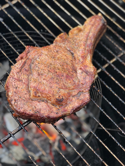 A close-up view of a large, grilled steak with a well-browned, seasoned crust, cooking over glowing charcoal on a barbecue grill.