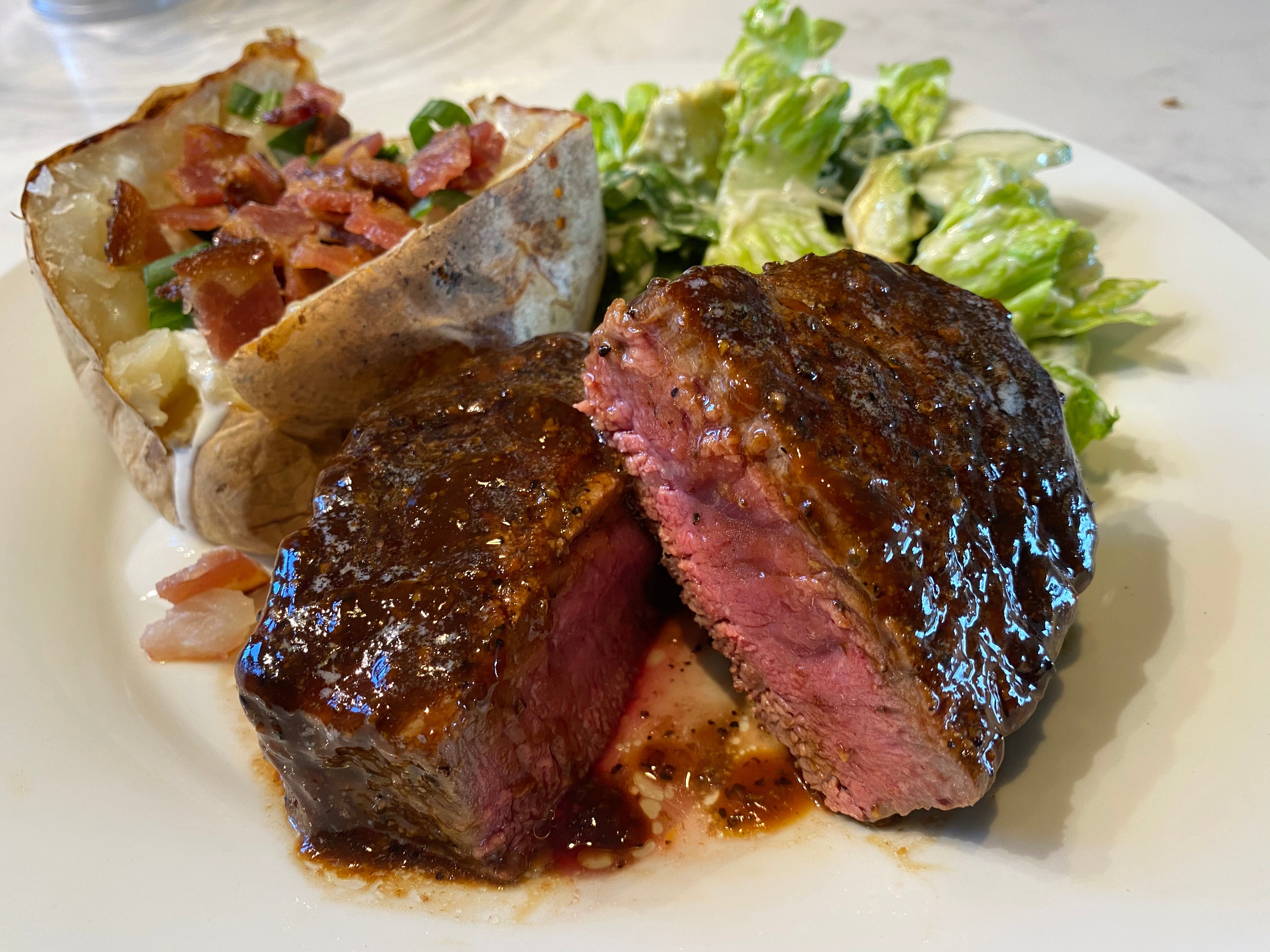 A succulent medium-rare steak, grilled to perfection with a glossy, caramelized exterior, served on a white plate alongside a baked potato topped with bacon bits and green onions, and a side of green leaf salad.
