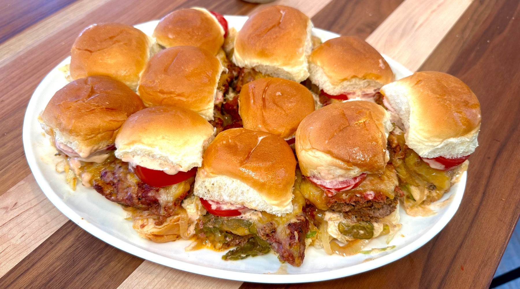 A plate of small sliders on soft, golden buns, filled with melted cheese, sliced tomatoes, and burger patties. The sliders are arranged closely together on a white plate placed on a wooden table.