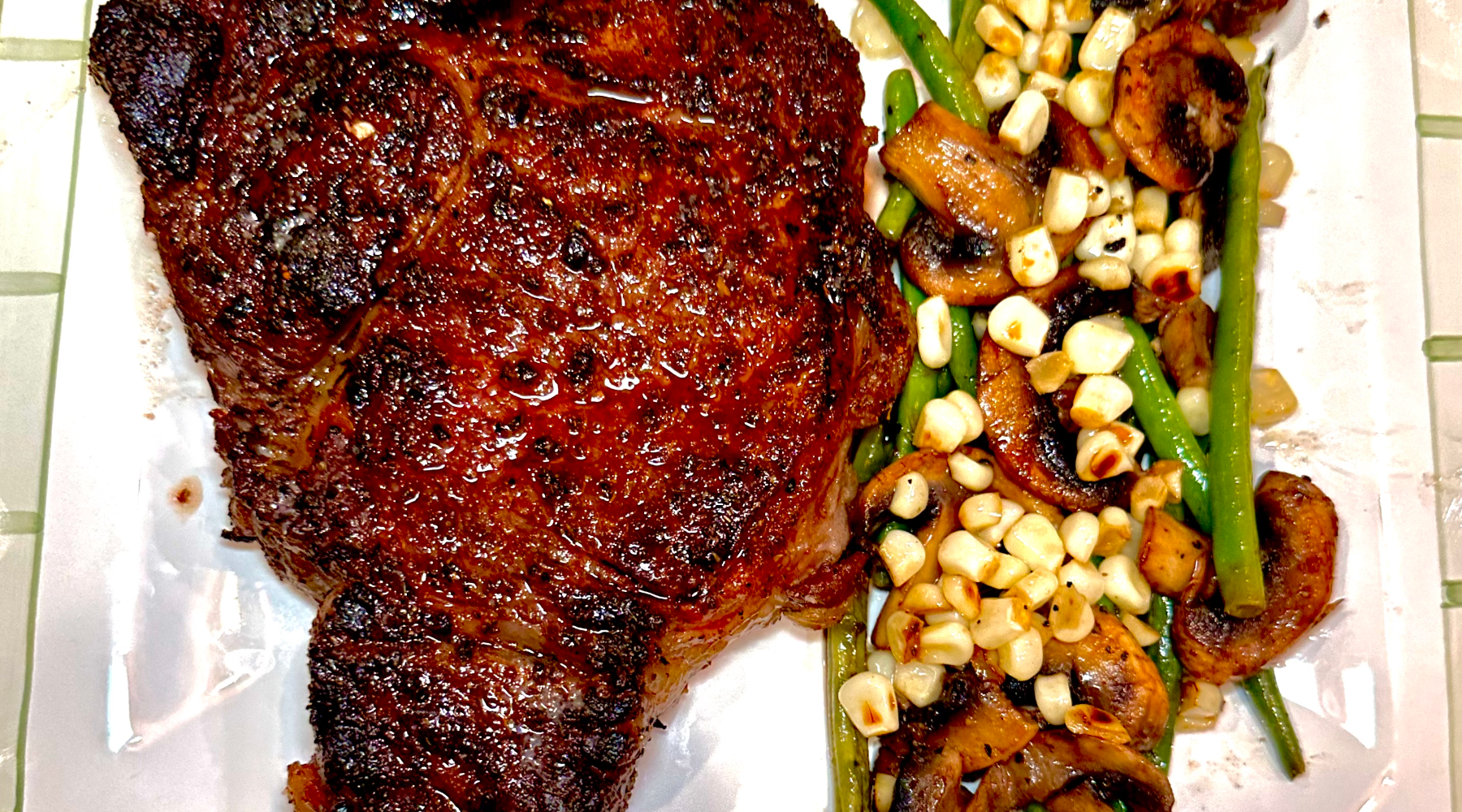 A grilled, well-seared steak served on a white plate alongside a medley of sautéed green beans, mushrooms, and corn kernels. The steak has a rich, caramelized crust, and the vegetables are lightly browned