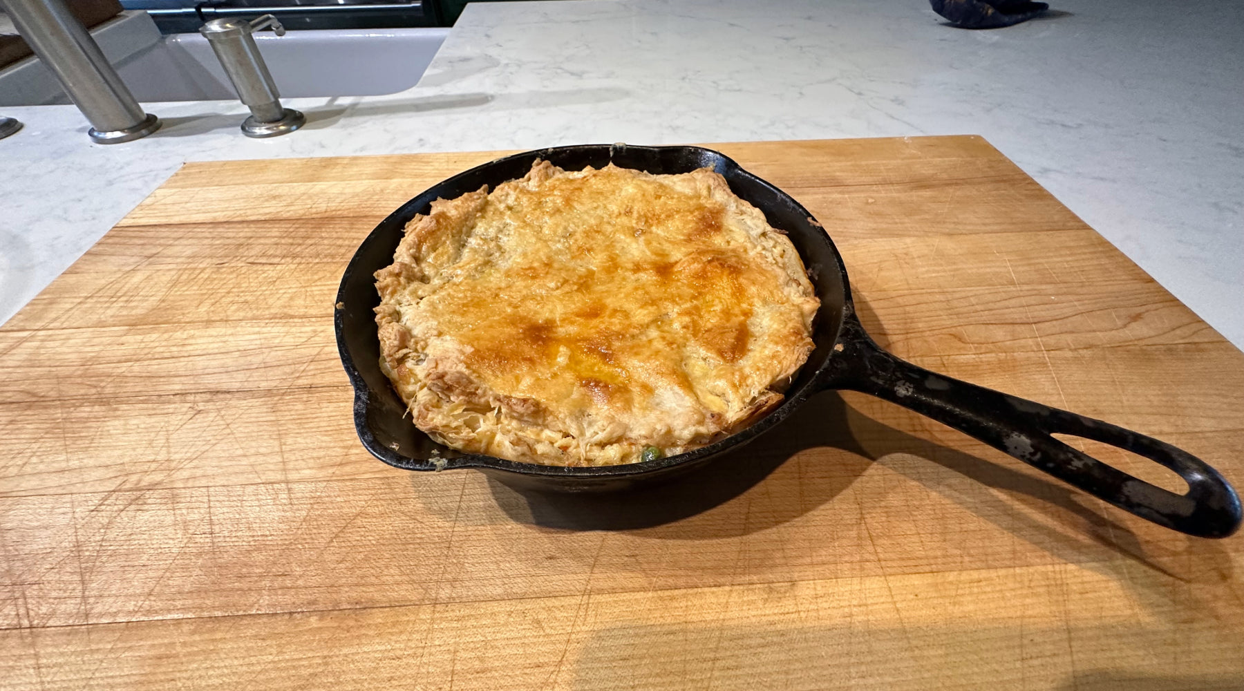 A golden-brown pot pie baked in a cast-iron skillet, resting on a wooden cutting board. The pie has a flaky, crusty top, and the setting appears to be a kitchen counter with a sink in the background.