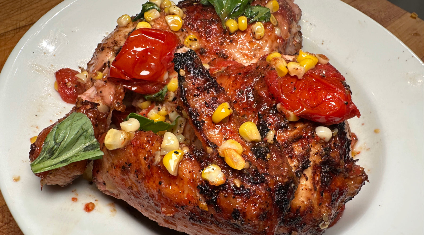 Grilled chicken with charred skin, topped with roasted cherry tomatoes, corn, and fresh basil on a white plate.