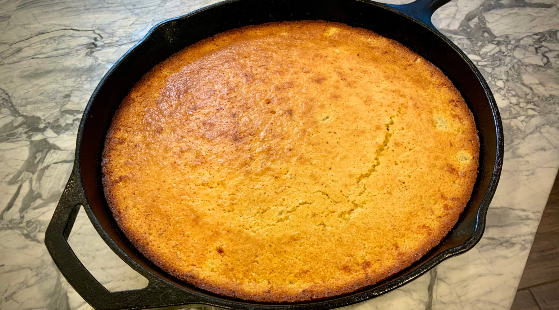 Golden-brown cornbread with a slightly cracked surface, freshly baked in a black cast-iron skillet on a marble countertop.