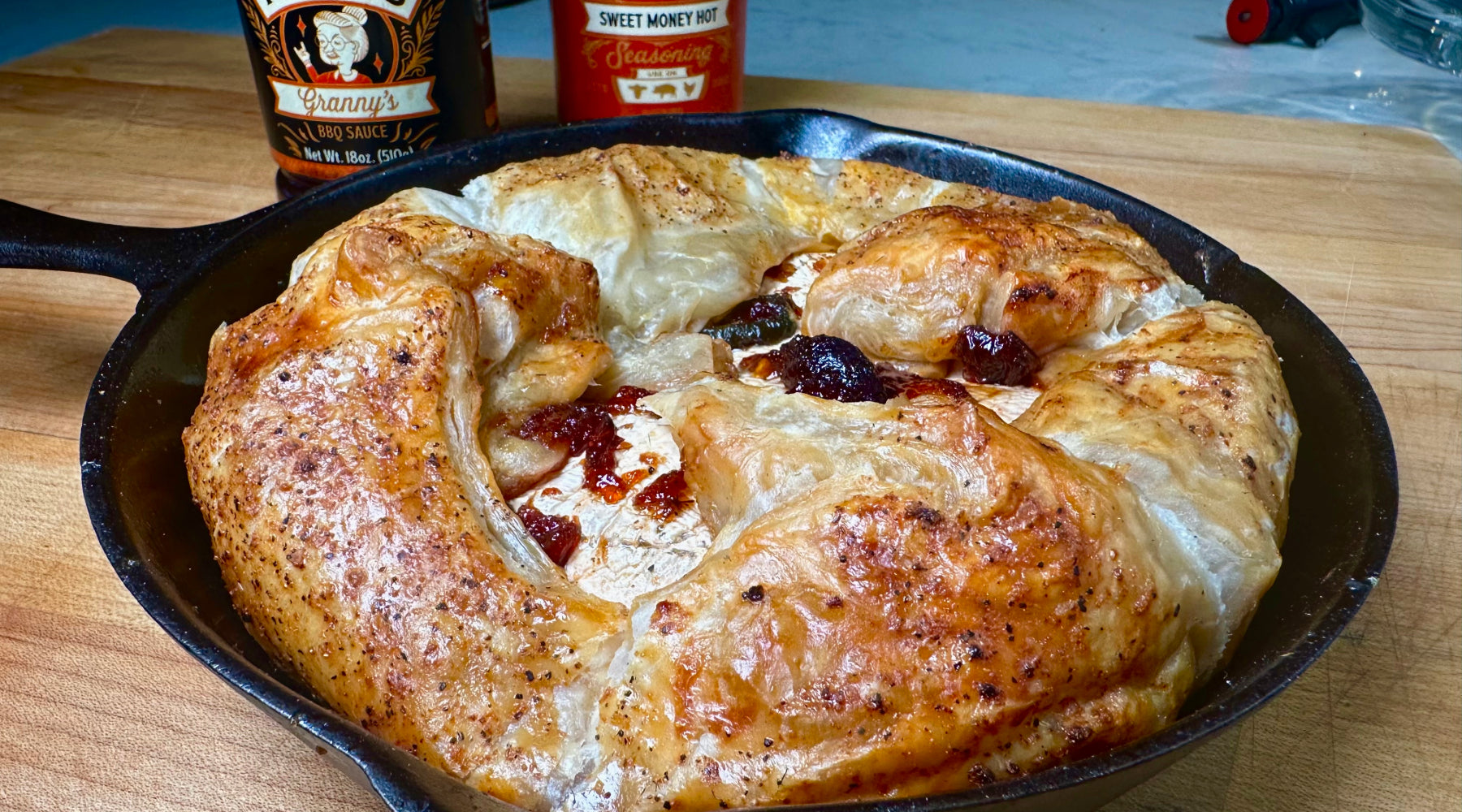 Baked brie in a skillet with flaky puff pastry, sun-dried tomatoes, and seasoning, placed on a wooden surface with sauce bottles nearby.