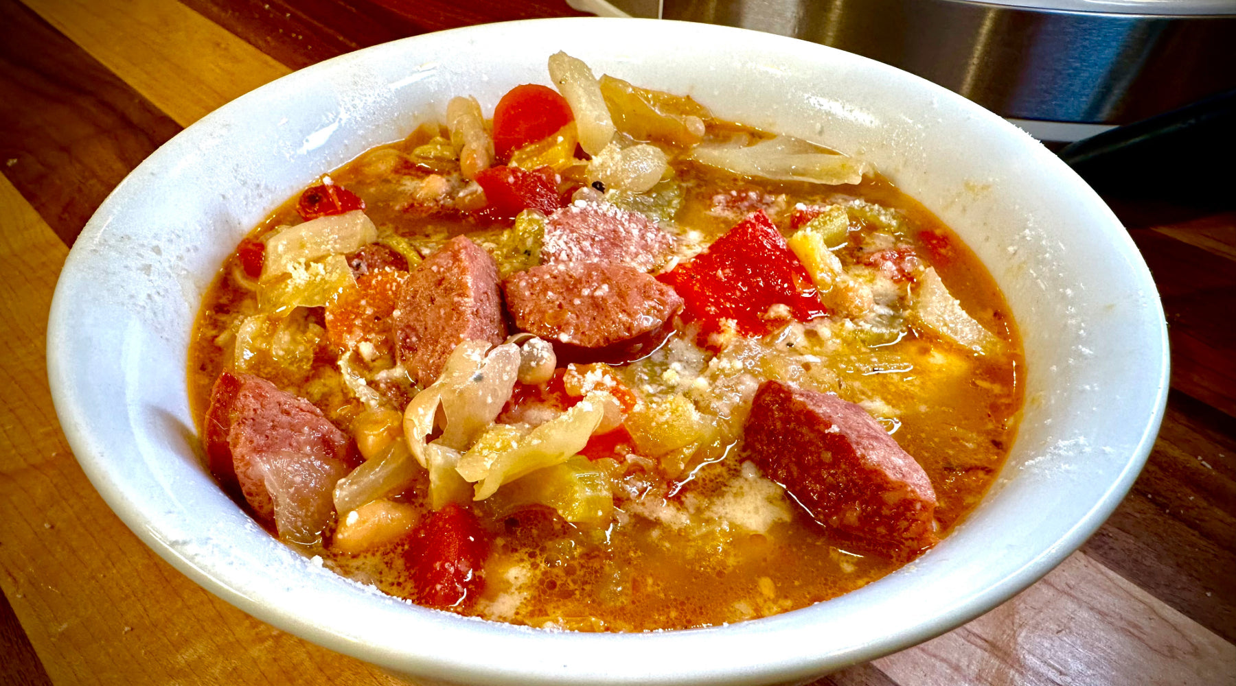 A bowl of sausage and vegetable soup with tomatoes, onions, beans, and broth, garnished with grated cheese, served on a wooden surface.