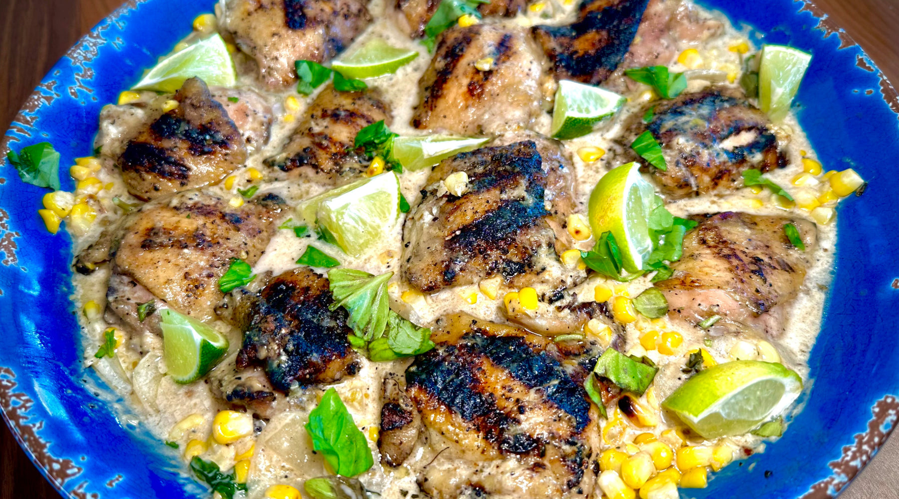 Grilled chicken thighs in a creamy sauce with corn, lime wedges, and basil, served on a blue plate.