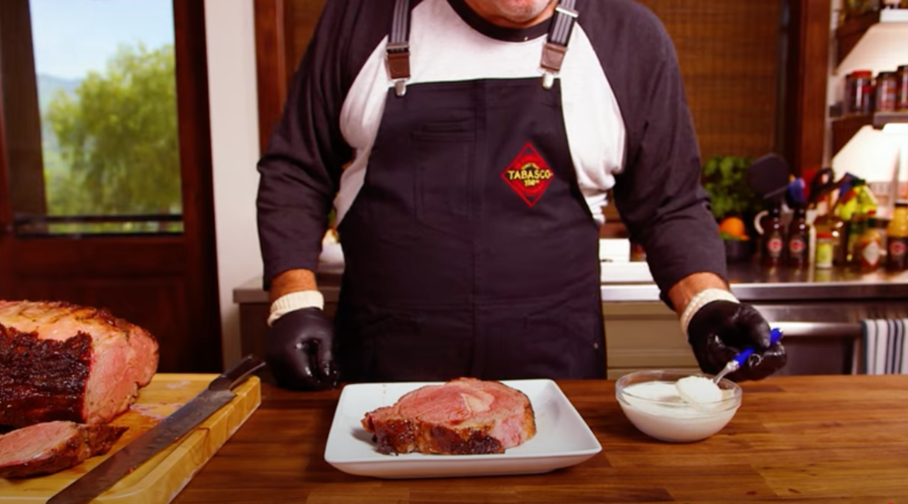 A chef in a black apron prepares a sliced prime rib with horseradish sauce in a kitchen.