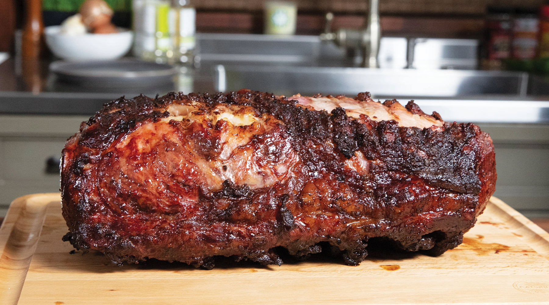 Juicy, smoked prime rib with a crispy crust resting on a wooden cutting board in a kitchen.